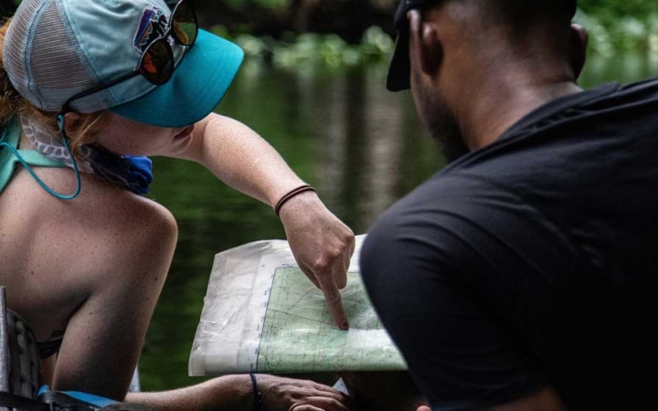 Two people who appear to be sitting in canoes examine a map. One person points at the map.